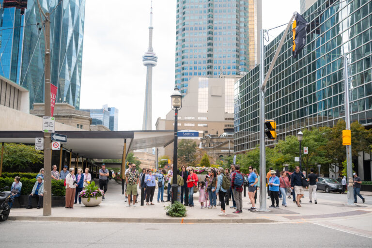 A group of people stand around a sidewalk with traffic lights and signs around. In the background is the CN Tower and skyscrapers prominently featured.