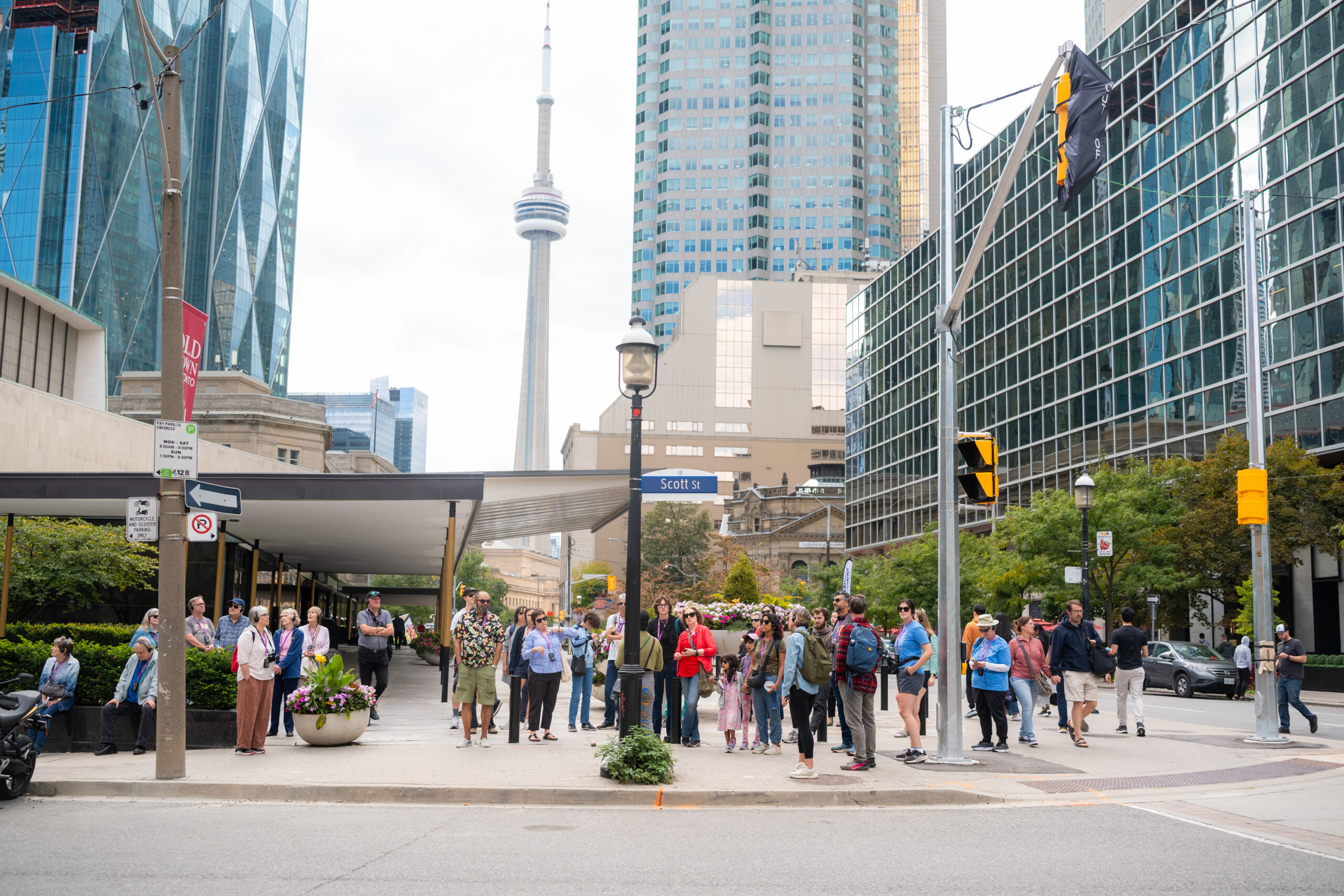 A group of people stand around a sidewalk with traffic lights and signs around. In the background is the CN Tower and skyscrapers prominently featured.