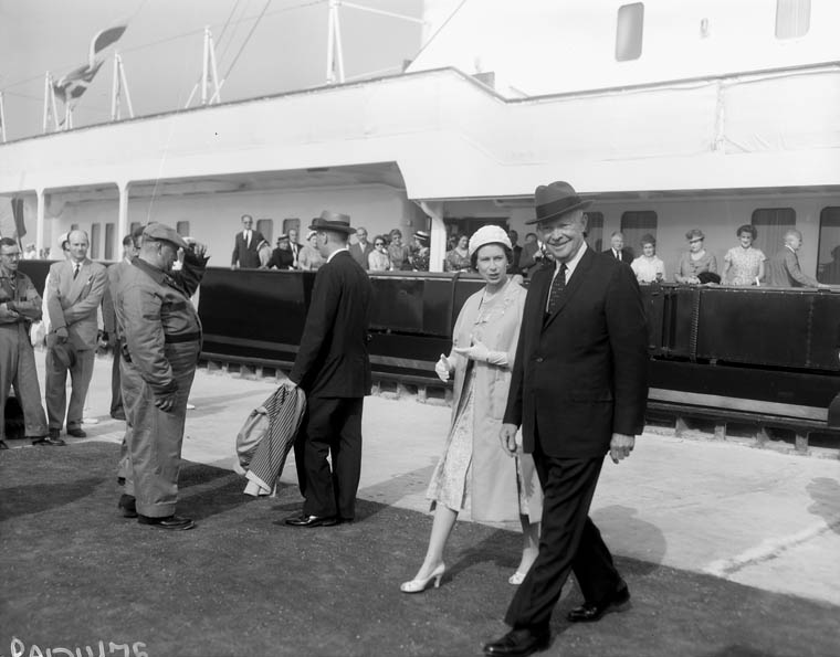 Black and white photograph of several people in front of a ship. Closest to the photographer, two people are strolling from left to right. A formally dressed man — with a suit, tie, and hat  — smiles at the camera. To his left is a woman in a long, light-colored jacket, wearing high heels, gloves, and a hat. She is saying something to the man. Behind them, a formally dressed man stands near another wearing overalls and a driving cap. The man is grabbing his hat, possibly greeting someone. The ship, at least two stories, extends out of view. Many men and women stand on the deck of the boat, looking at the man and woman at the front of the image. 