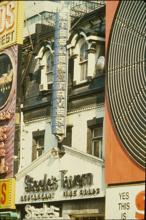 A colourful photograph depicting a bulb vertical sign on a white brick building with a black roof. The sign is blue and reads "Steeles Tavern".