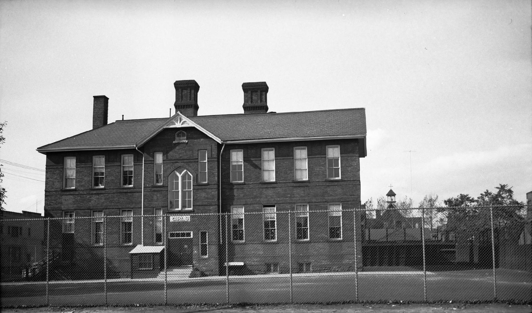 A black and white photo of a two-storey brick building with three chimneys. In the foreground is a grassy area.