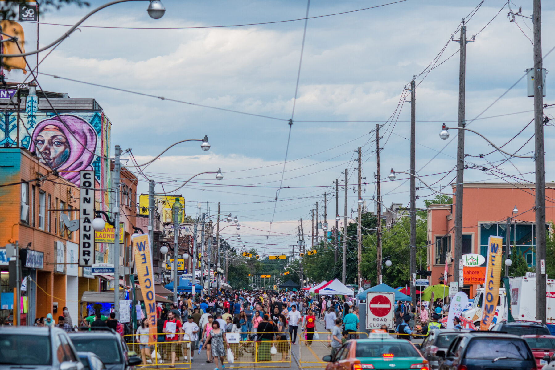A large crowd of people at a street festival. The street is closed for the event, and many signs and tents are displayed.