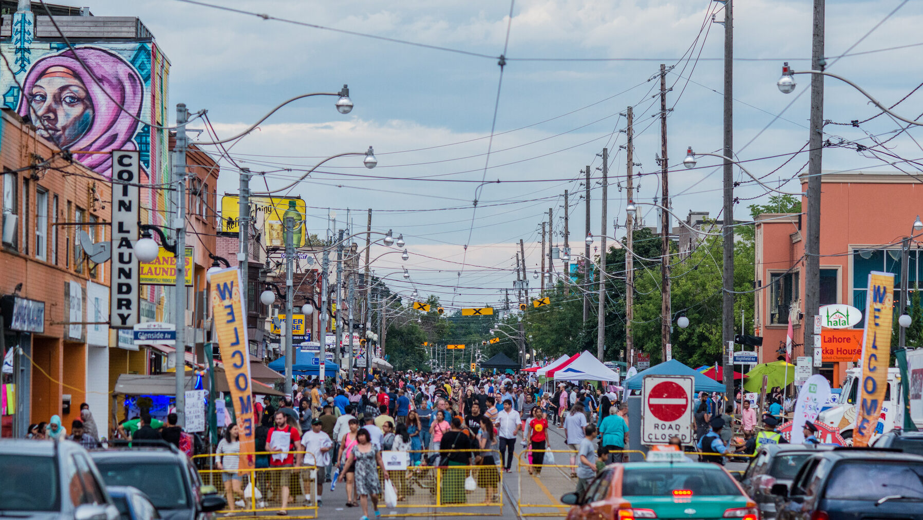 A large crowd of people at a street festival. The street is closed for the event, and many signs and tents are displayed.