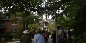 People are looking at a house on a residential street with large trees.