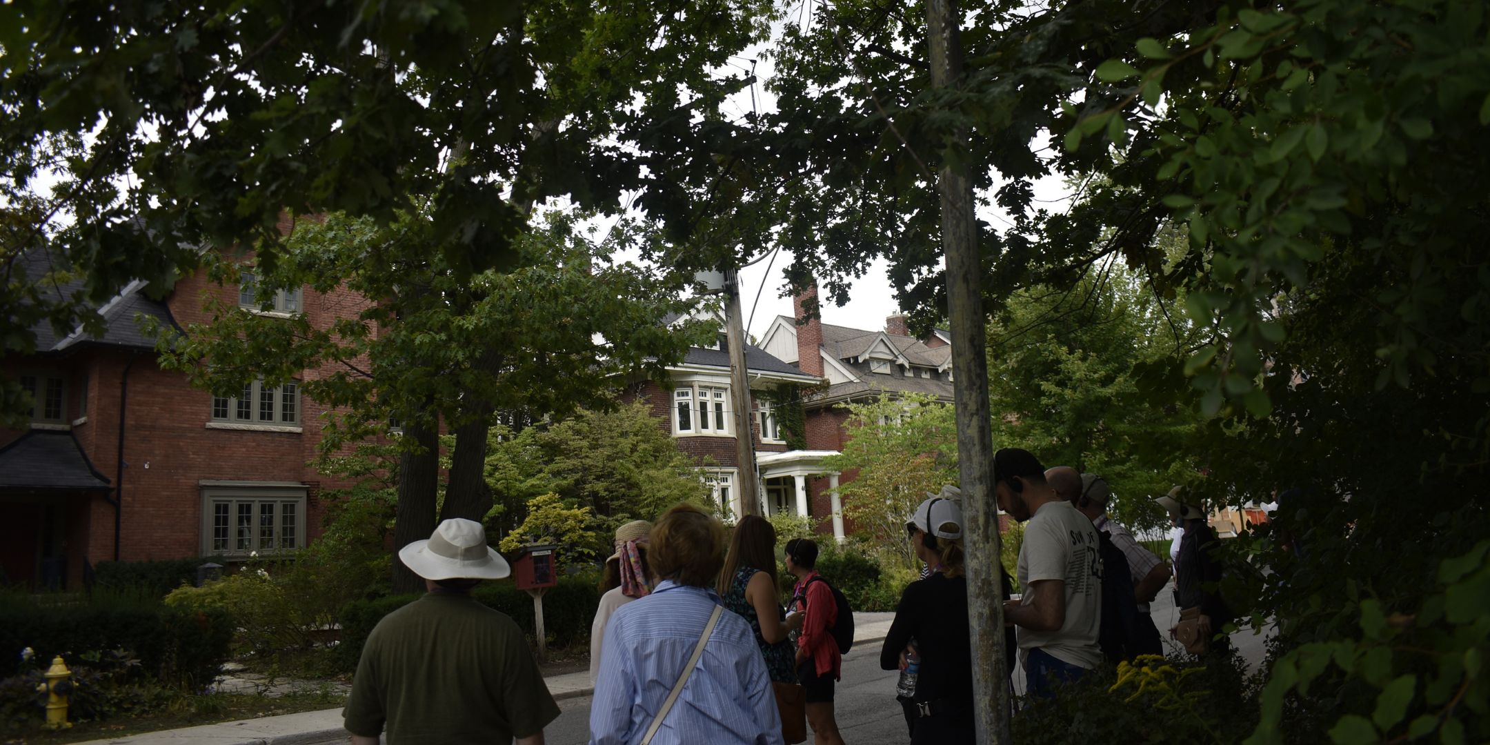 People are looking at a house on a residential street with large trees.