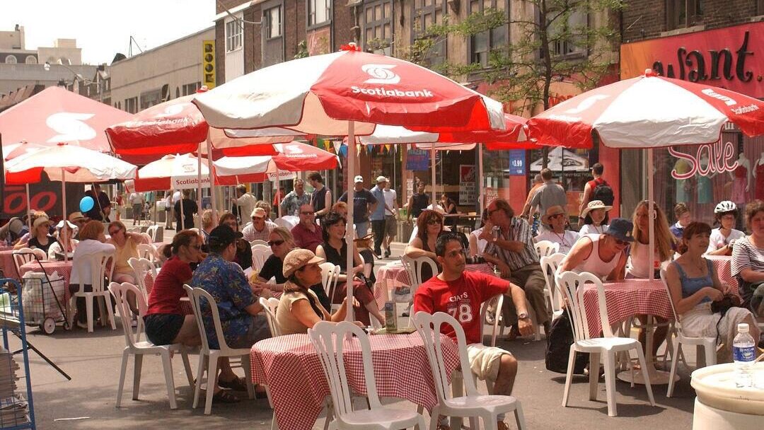 A crowd of people sit at round tables underneath red and white umbrellas.