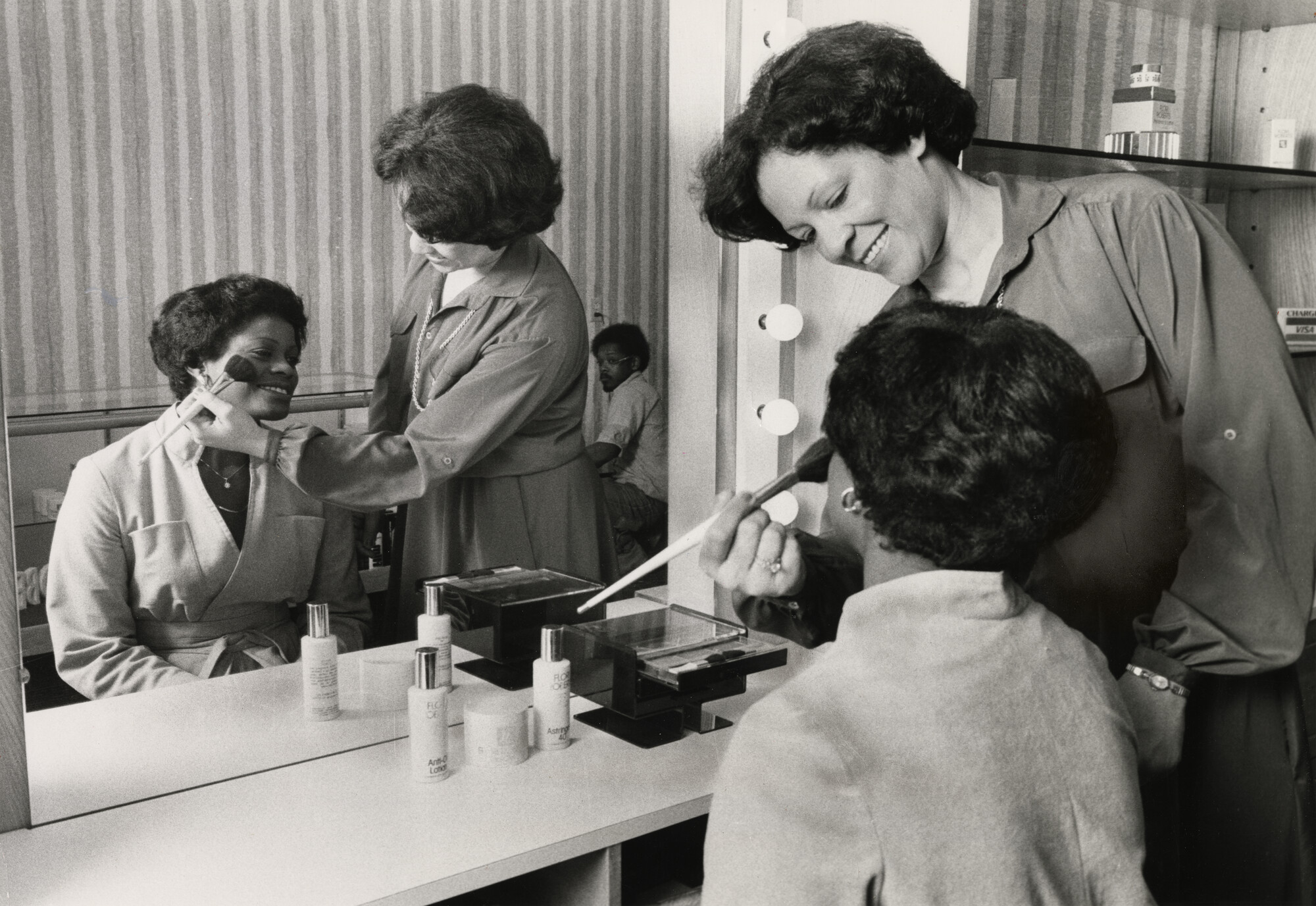A woman applies makeup to the cheek of another woman seated in front of a mirror.