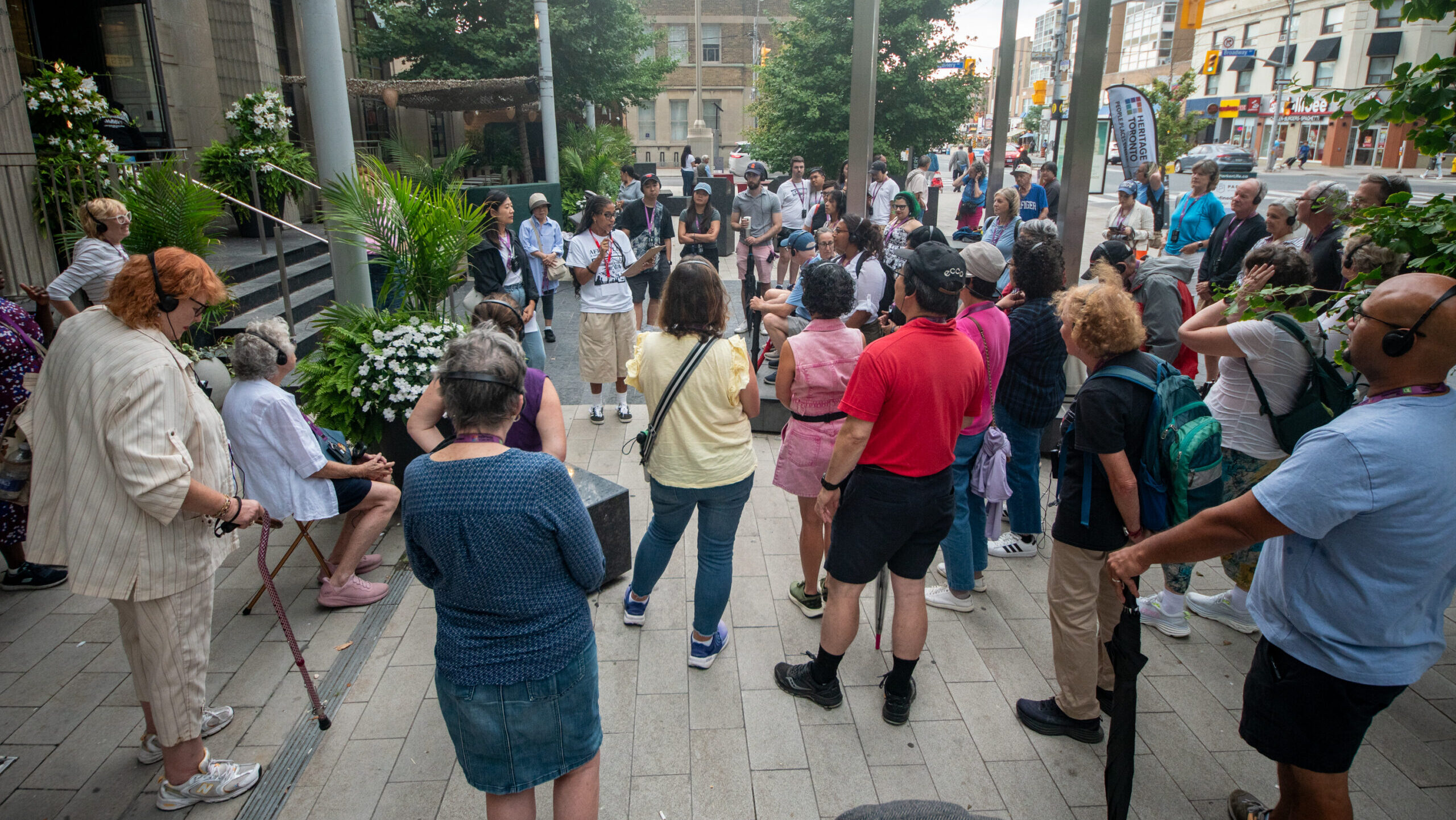 A group of people stand around on a paved grey and beige area with metallic structures erected around them, buildings in the background, and listen to someone in the center of the group speaking to them.