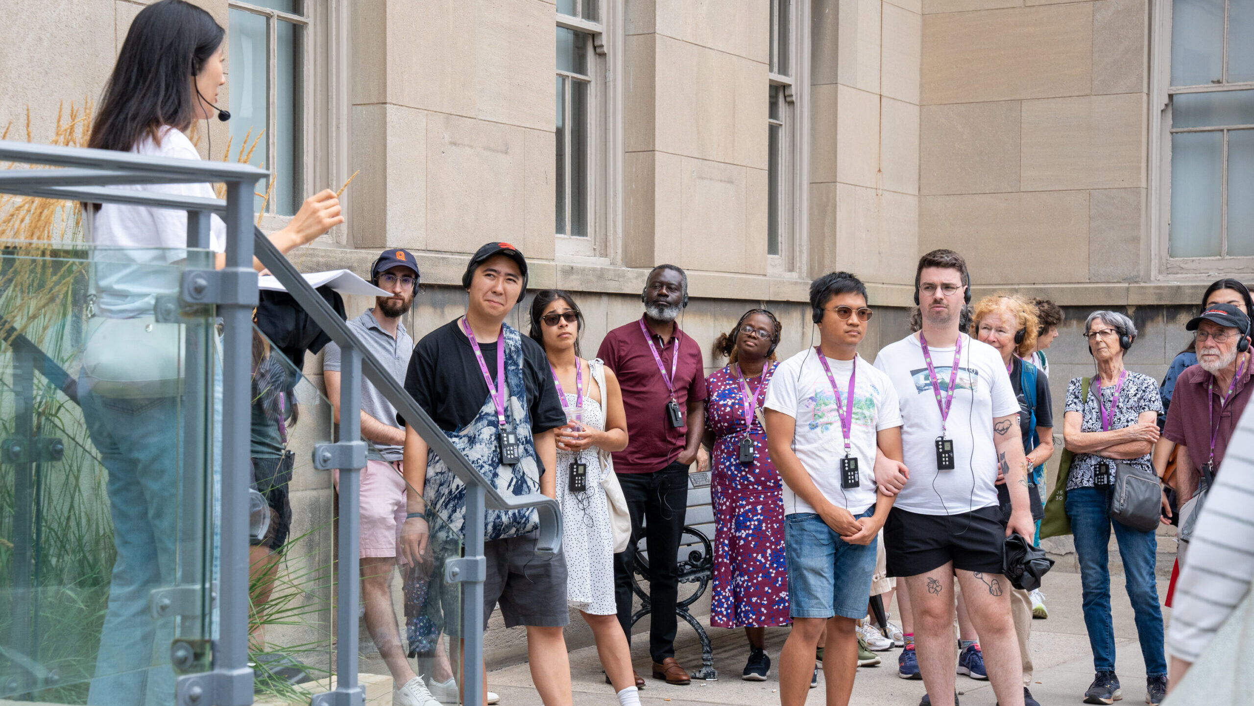 A group of people stand on a sidewalk in front of a beige building and look toward a person standing on a set of stairs near them, who is speaking to the group.