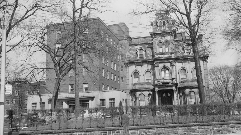 A black and white image of a hospital from the opposite side of the road. The property is fenced off and the yard is scattered with barren trees. The hospital has an older wing and a newer looking addition on its right side.