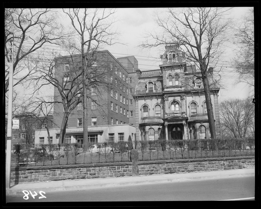 A black and white image of a hospital from the opposite side of the road. The property is fenced off and the yard is scattered with barren trees. The hospital has an older wing and a newer looking addition on its right side.