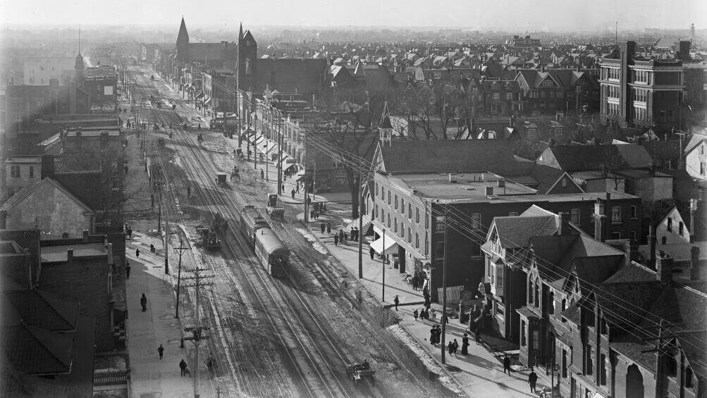 View of a road and surrounding buildings from a high vantage point. Old streetcars, horse drawn carriages, and pedestrians are depicted.