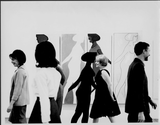 A black and white image of several people walking in various directions in front of a large piece of art. The art depicts the repeated figures of a naked woman, with various shading.
