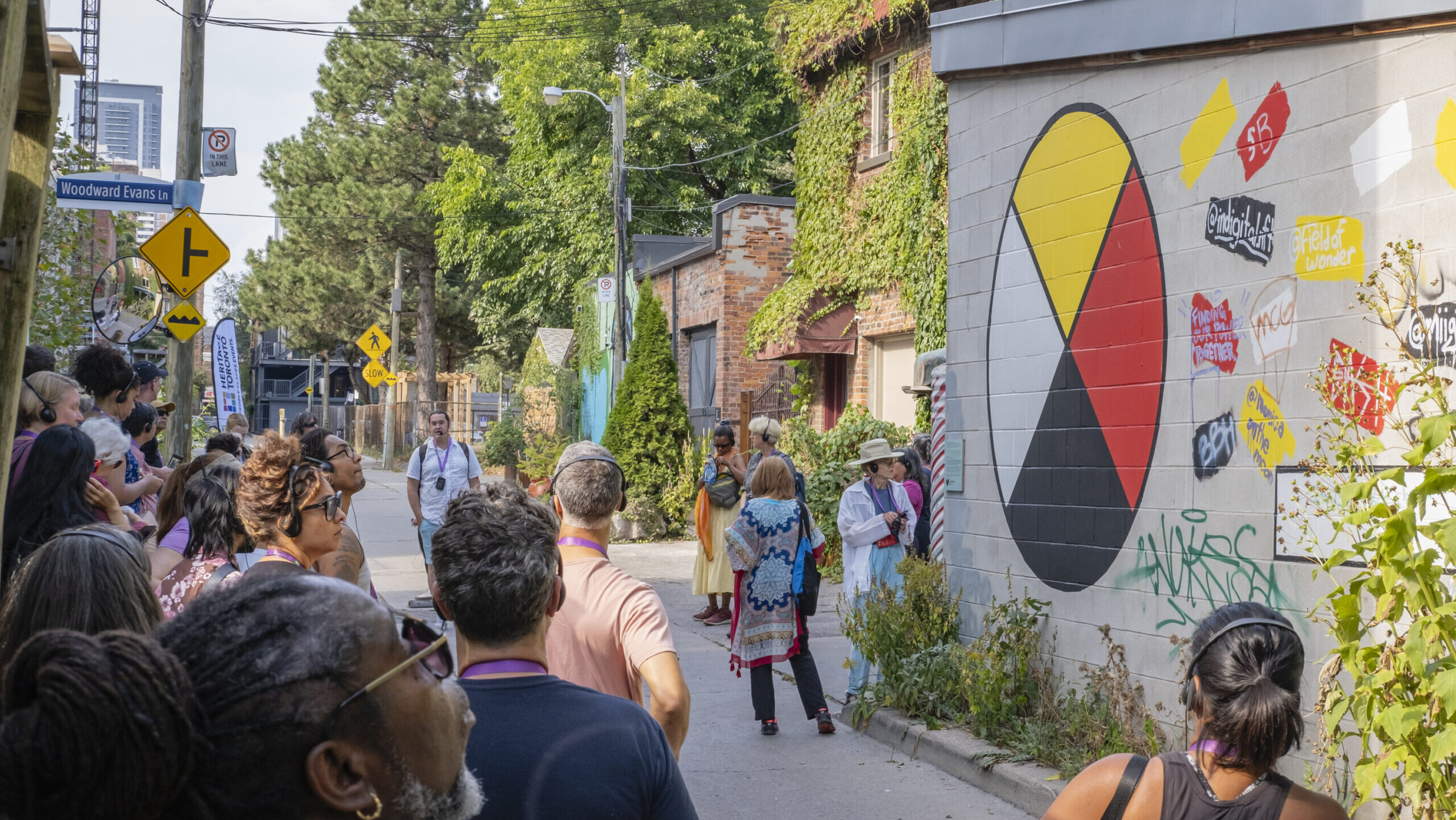 A group of people are standing in a laneway. On the wall of the building to the right of the laneway is a mural of an Indigenous medicine wheel (a circle with yellow, white, red, and black wedges). Large green trees are in the background.