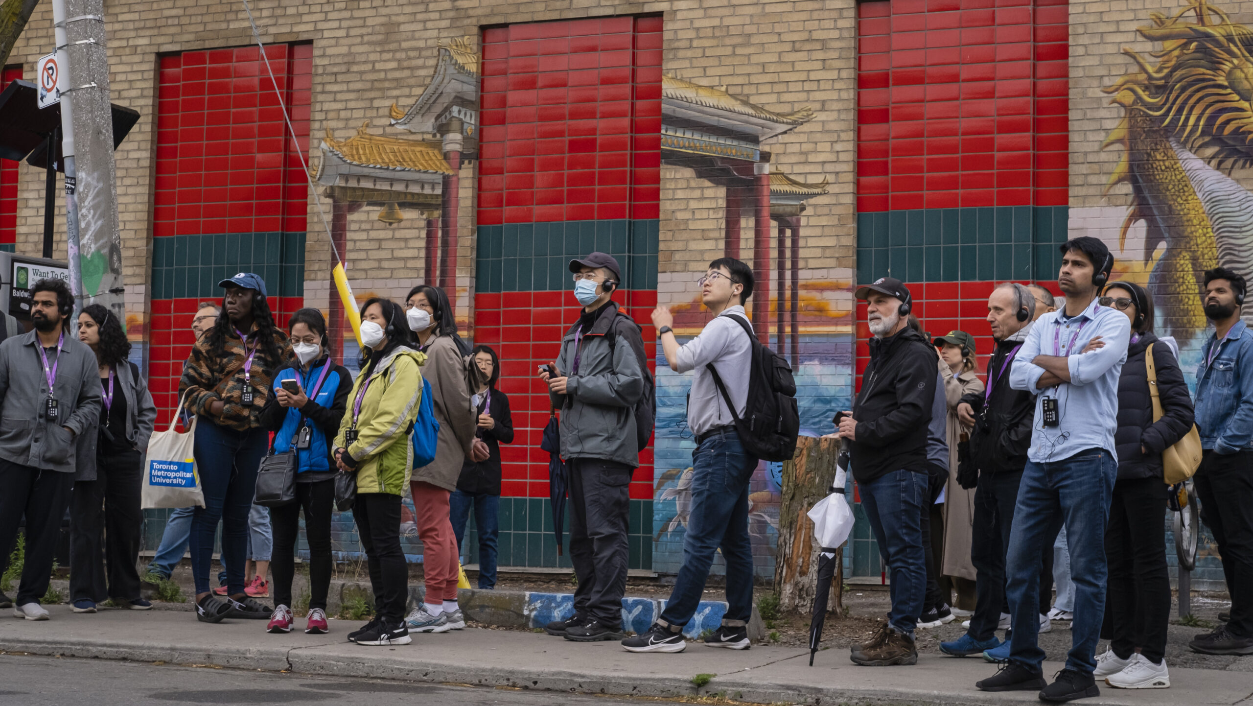 Group of people stand on a sidewalk against a brick building. It's exterior is brightly painted.