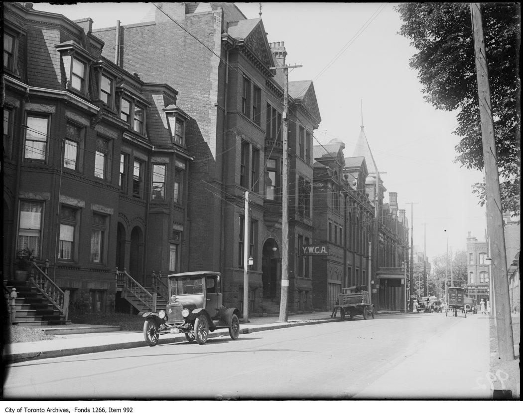 A quiet street with old-fashioned cars parked alongside it. Several brick buildings are on view, including one with a prominent sign that reads "YWCA".