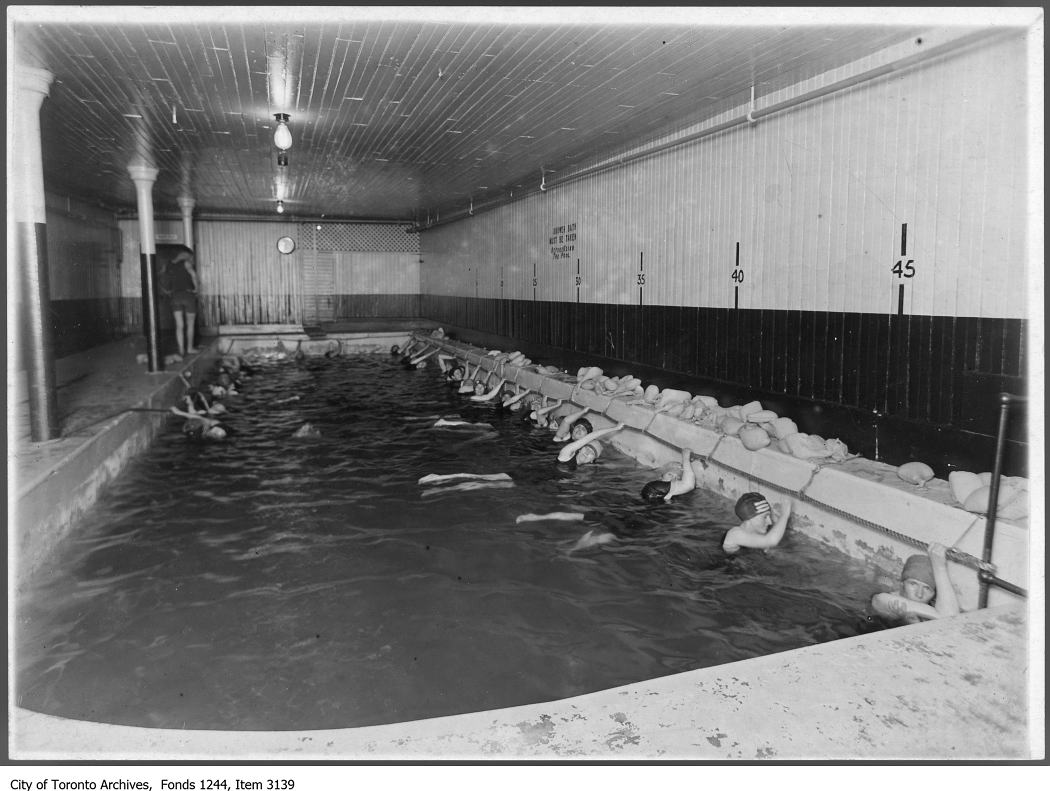 A small indoor pool is full of swimmers, all women and all holding on to the pool's sides. All wear swim caps and long swimwear.