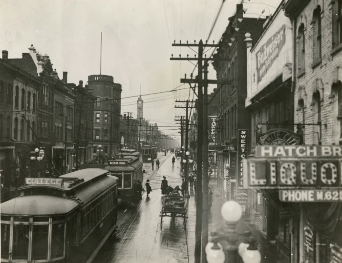 Black and white image of Yonge St from the vantage point of looking north from Granby St. Buildings of various architectural styles and electrical poles line the streets. Several of the buildings have electric signs hanging from the storefronts. Several streetcars are on the road, along with a horse-drawn carriage.