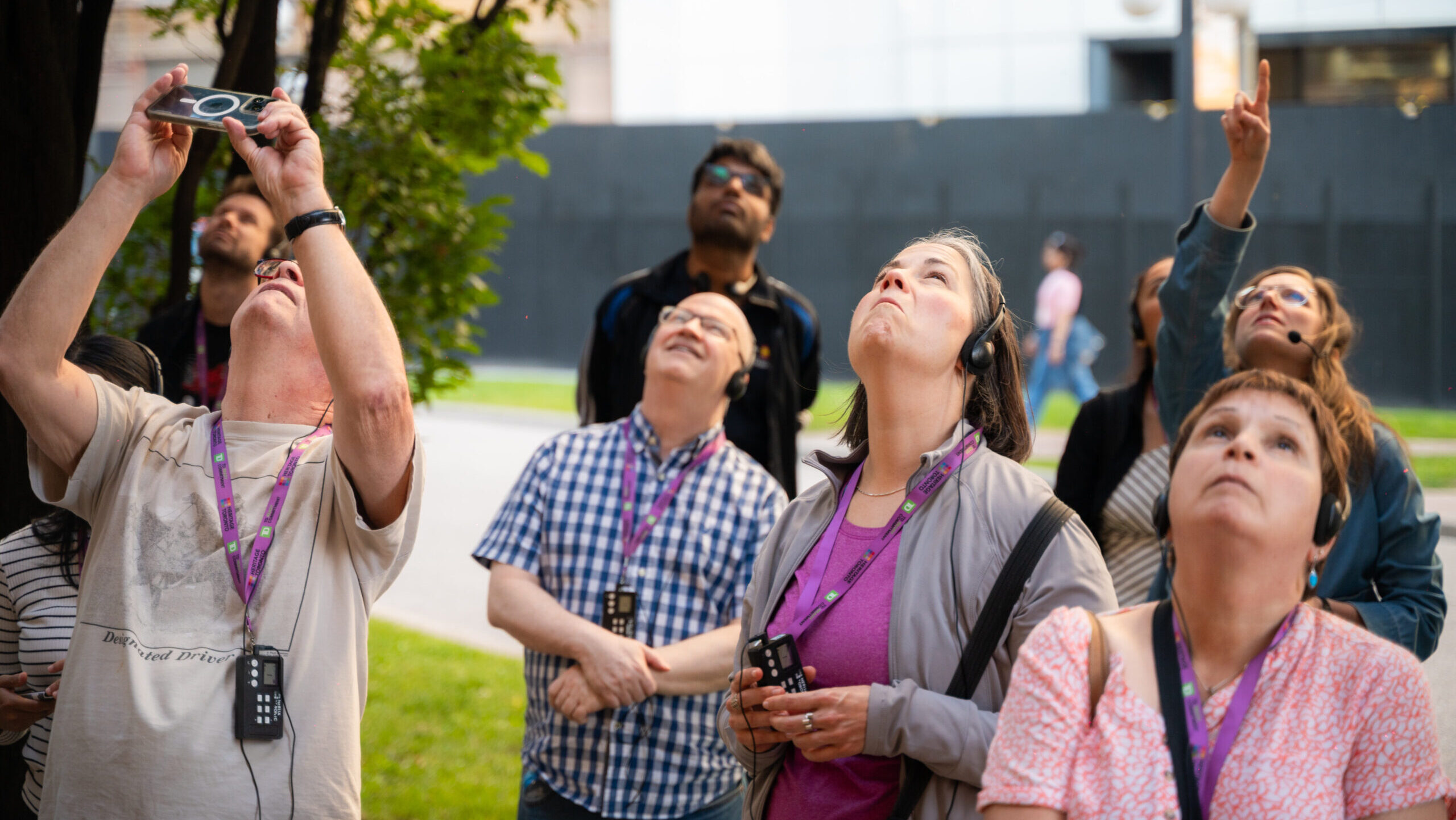 About eight people stand and look up. They wear headsets and one is pointing with her hand while another takes a photo with their phone.