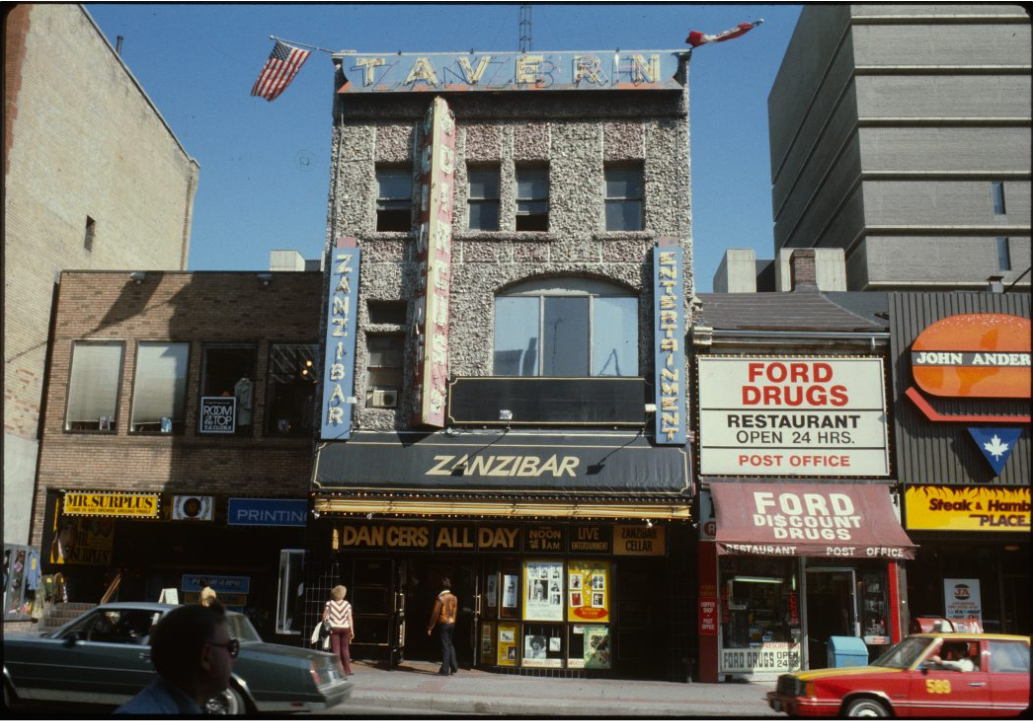 A colour photograph portrays a three story rectangular building with a sign at the top that reads "Tavern". Cars drive by on the road in front. There are neon signs in the window.