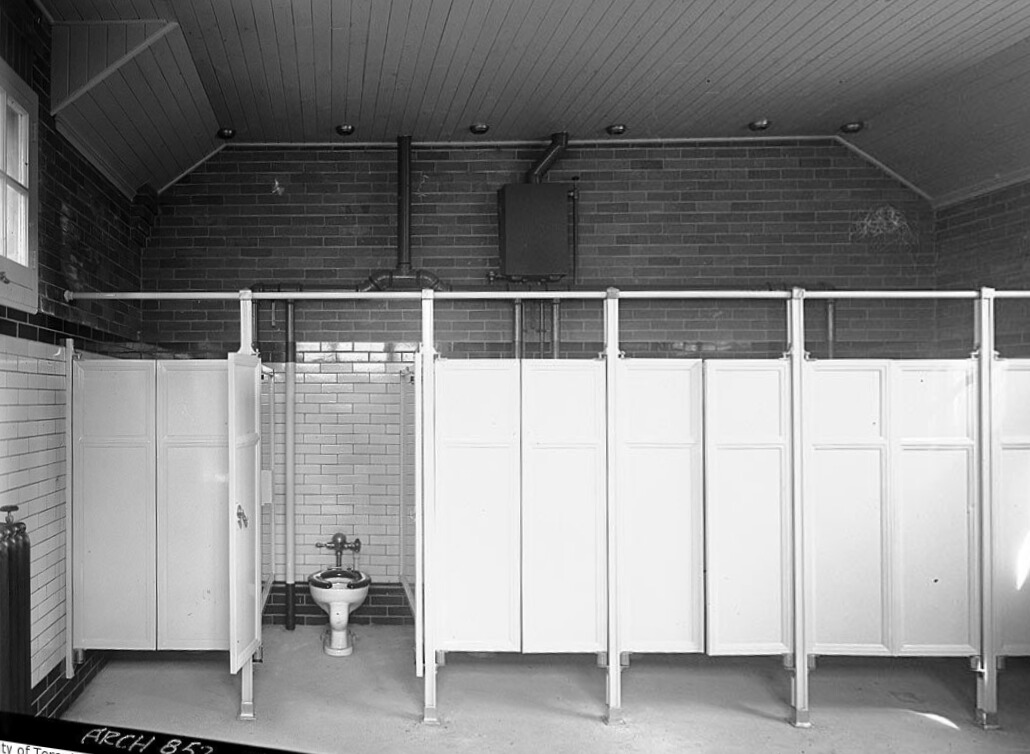 Black and white image of six washroom stalls with one having an open door showing a lidless-toilet.