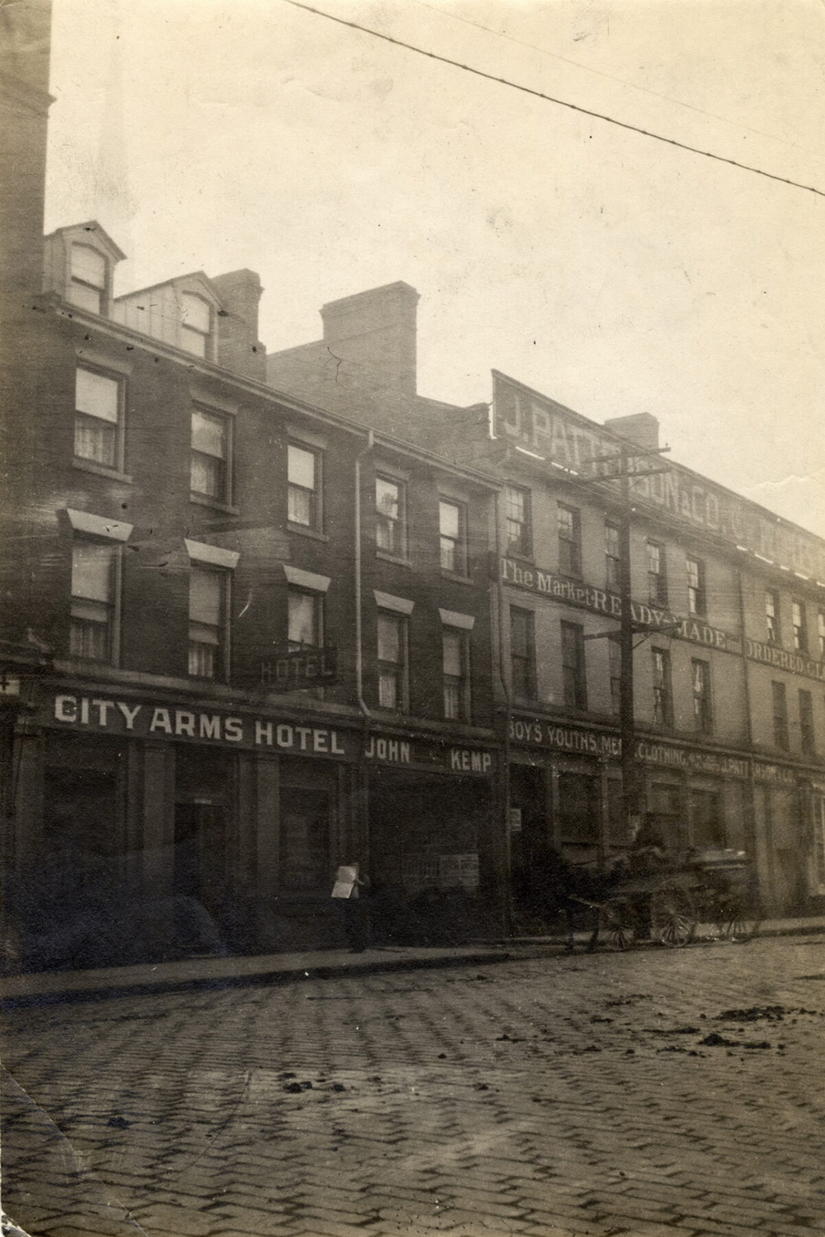 Black and white photo of a cobblestone street with buildings alongside it. One building's sign reads, "City Arm Hotel" and beside that, "John Kemp". The sign on the building beside this reads, "Patterson & Co." and various other shop signs are visible on the same building. A horse-drawn carriage is in front of the shops on the road.