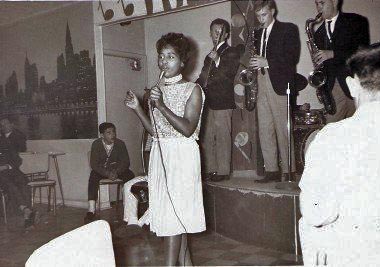 A black and white image of a female singer with dark skin singing into a handheld microphone, wearing a sequined light dress. Behind her on a stage stands a guitarist and two saxophone players, all men wearing suit jackets.