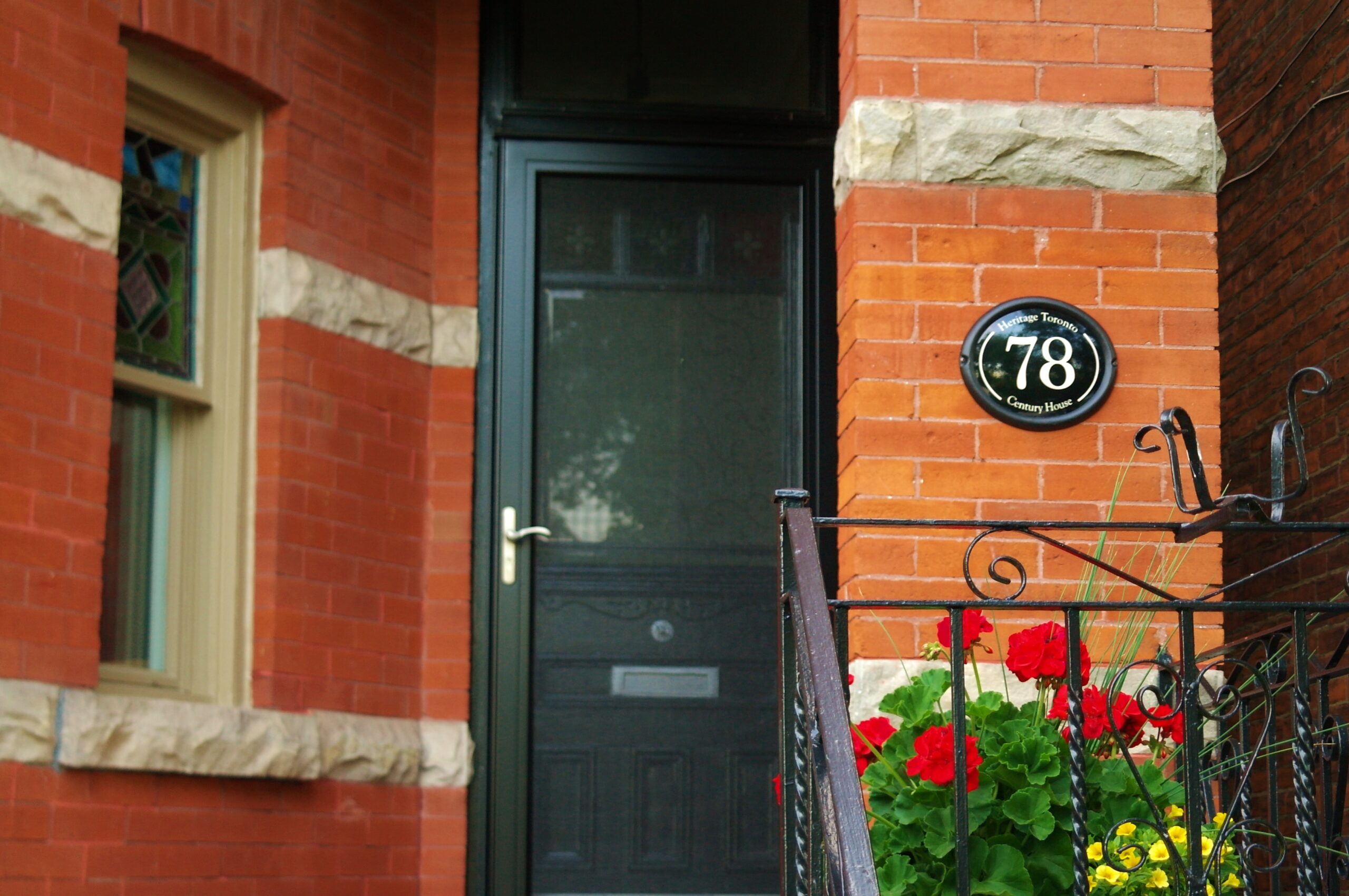 Image of a plaque mounted on a red brick wall on a porch. A railing, front door, window, and flower pot planted with red geraniums are all visible. The plaque is oval in shape, with a black background and text that reads 78.