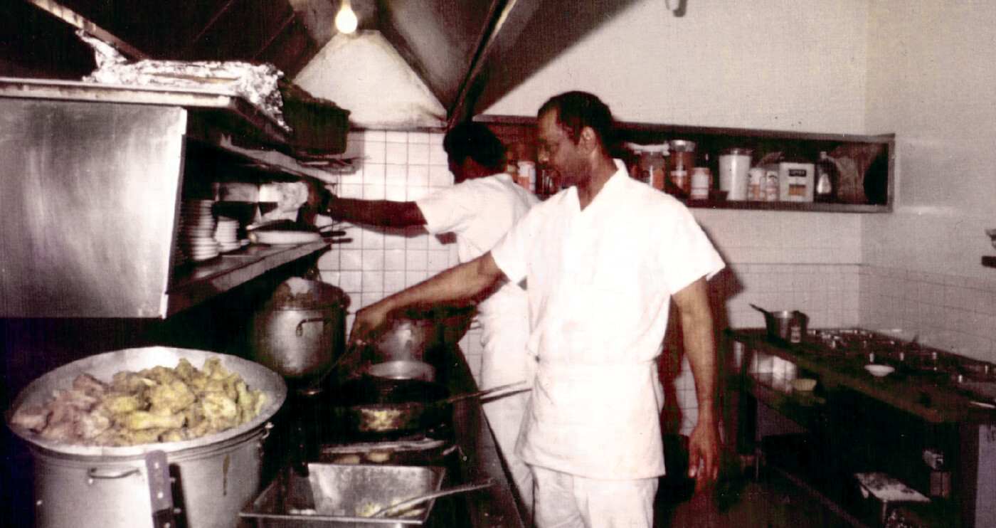 Image of two chefs in a small kitchen preparing food in front of a stove.