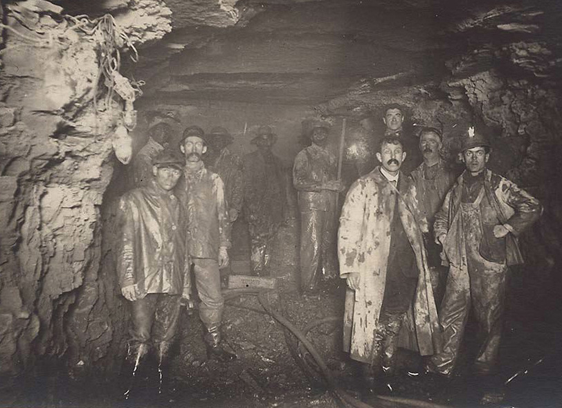 A black and white photograph of a group of men working in an underground tunnel. Their clothes are filled with mud and dirt.