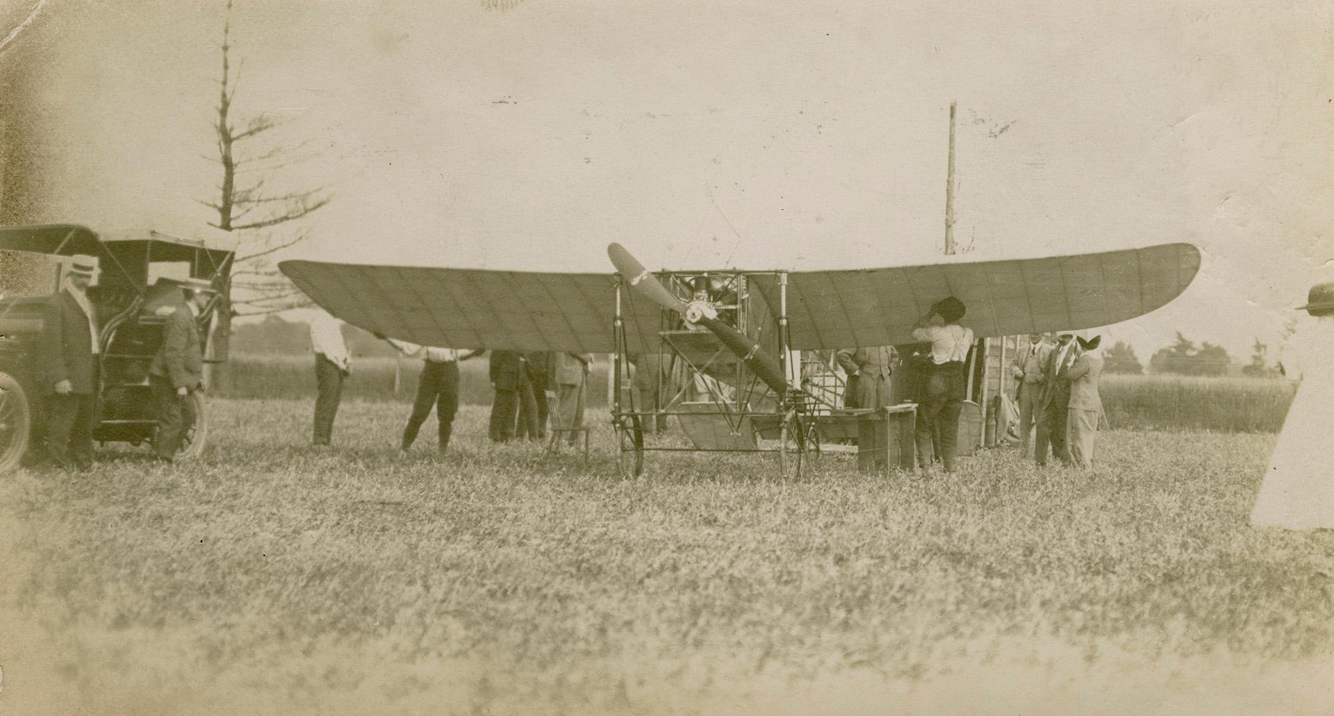 Sepia photograph of people standing around an early-style airplane in an open field. The plane's wings are blocking the people standing around it, so only their legs are in the image.