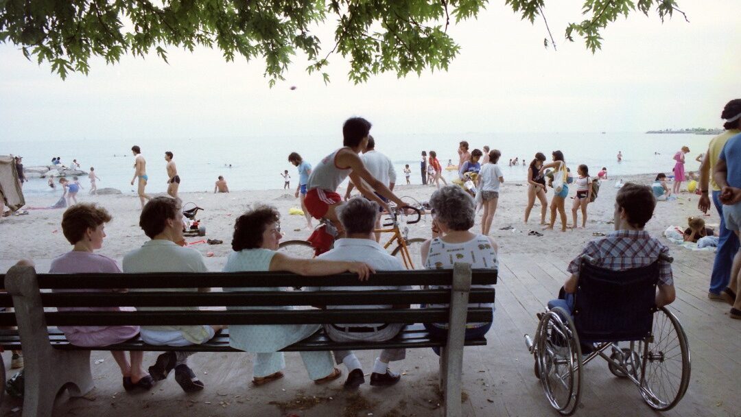 Five people sitting on a bench and one person sitting in a wheelchair facing a beach and boardwalk. Crowds of people are variously cycling, walking, and swimming.