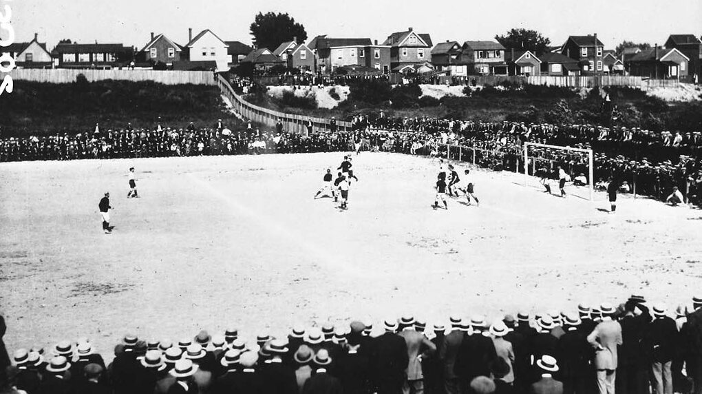 Wide view of a soccer game in progress. A large crowd of spectators are watching the game, standing behind the field boundaries.
