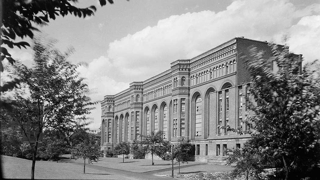 Image of a multi-storey building with large sweeping arched windows set along a tree lined avenue.