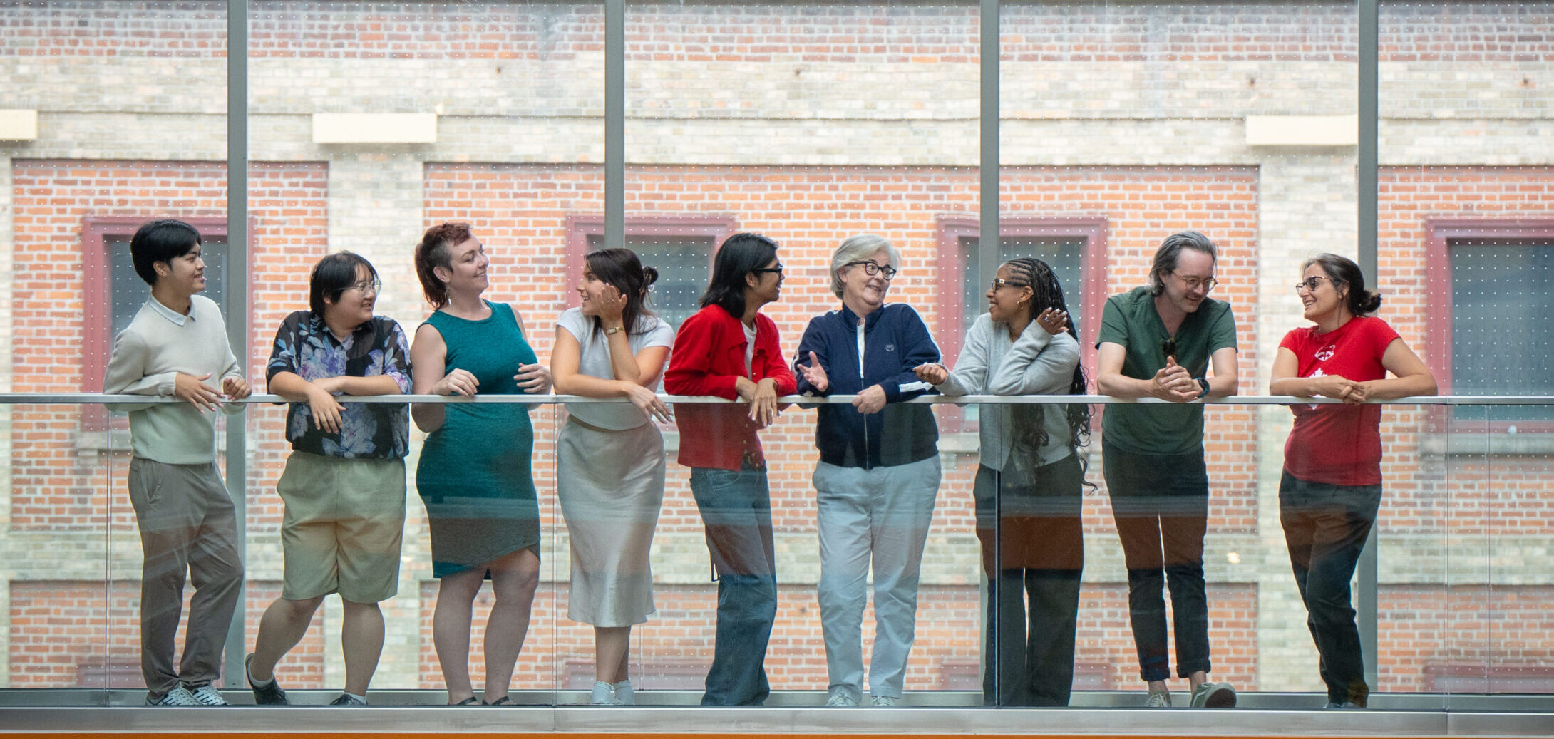Photo of nine people smiling and talking to each other behind a glass railing. Behind them are large glass windows through which a brick building is visible with the sign ST LAWRENCE MARKET