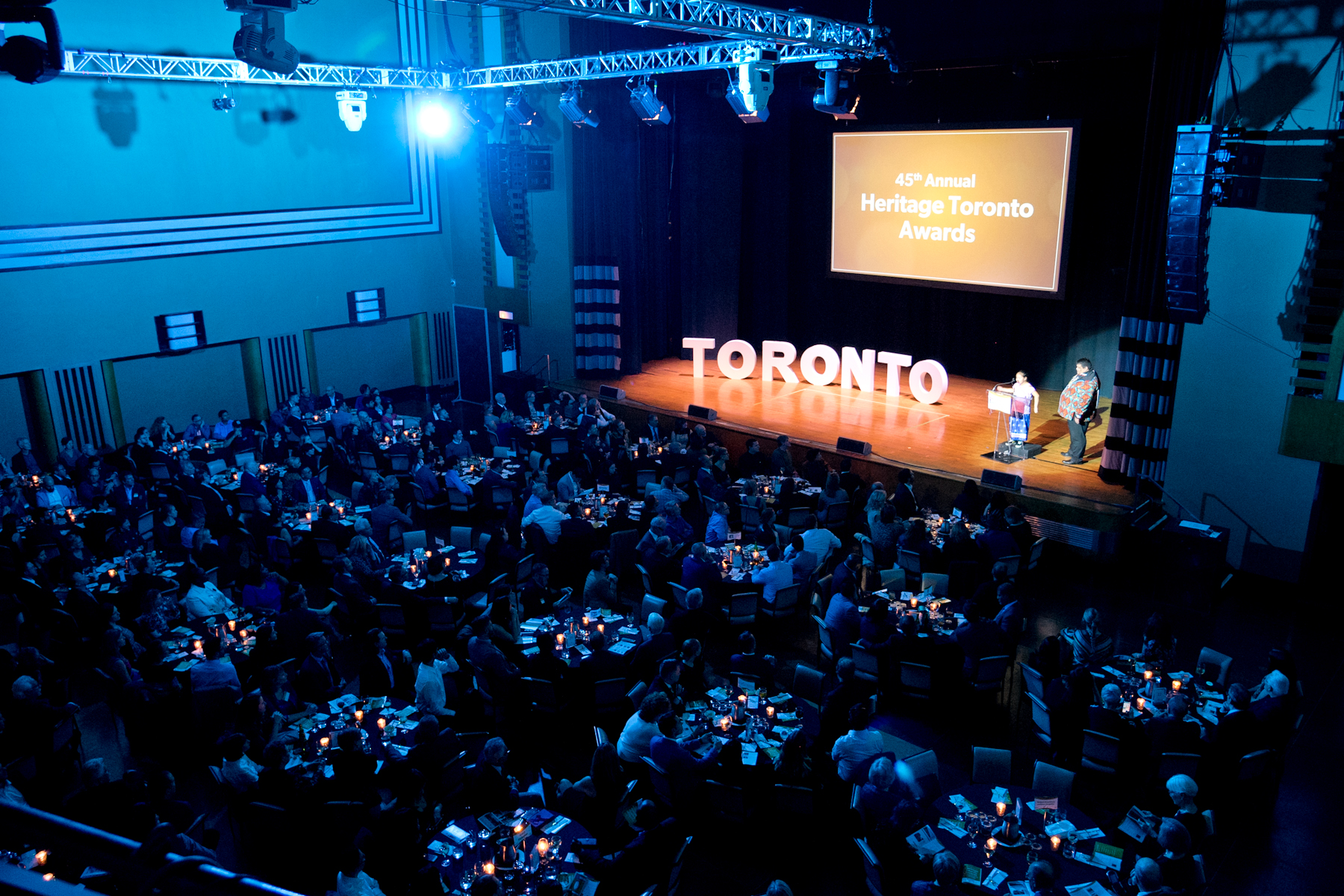 A photo taken from above looking down on a stage with a man and girl standing at the podium and large white letters spelling out Toronto to his left. Above and behind is a screen projection with the words 45th Annual Heritage Toronto Awards. On the floor of the auditorium are groups of people sitting at round tables.