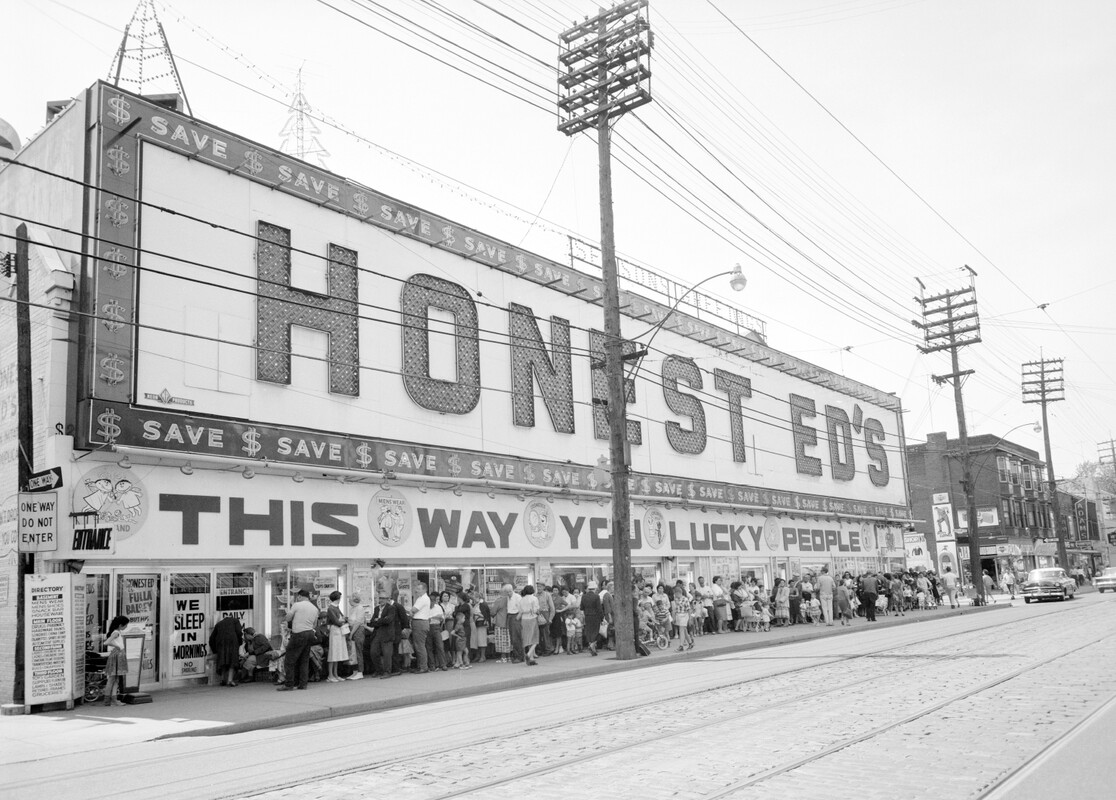 Black and white photo of the exterior of a large store with a gigantic sign that reads: "Honest Ed's - This Way You Lucky People".