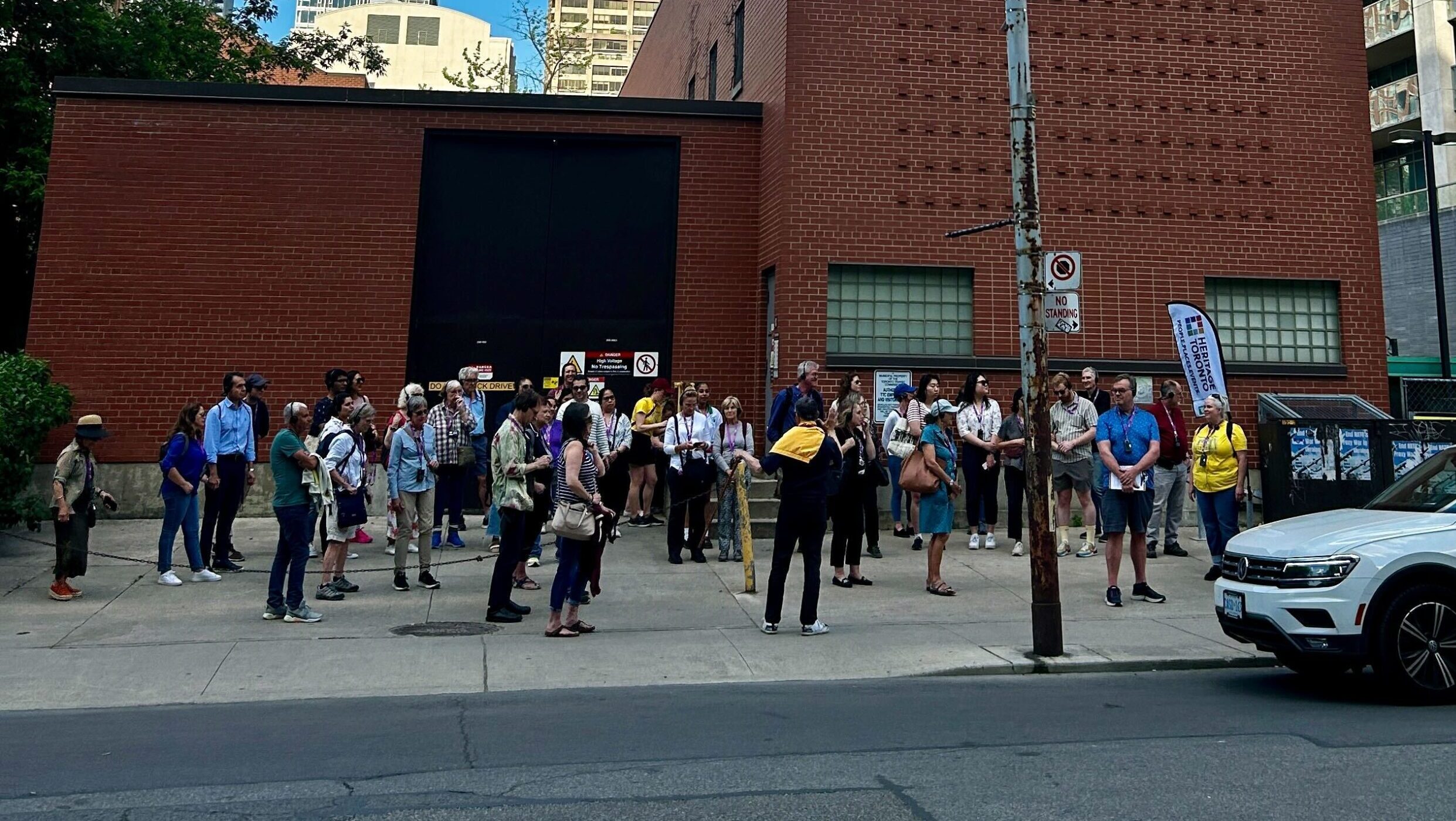 A large tour group is gathered on the sidewalk in front of a brown building on the U of T campus. The left side of the building is a single story, with a large black door. The right side of the building is two stories with two small rectangular windows on the lower story and one long rectangular window at the top of the building. The Heritage Toronto flag is located on the right side of the tour group.