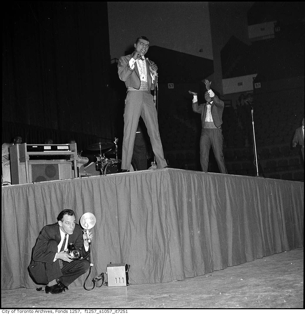 A black and white photograph of two men wearing stylized tuxedos singing on stage.  A man in a suit holding a camera in one hand and large flash bulb in the other is crouched down next to the stage, in the bottom left-hand corner of the photograph.