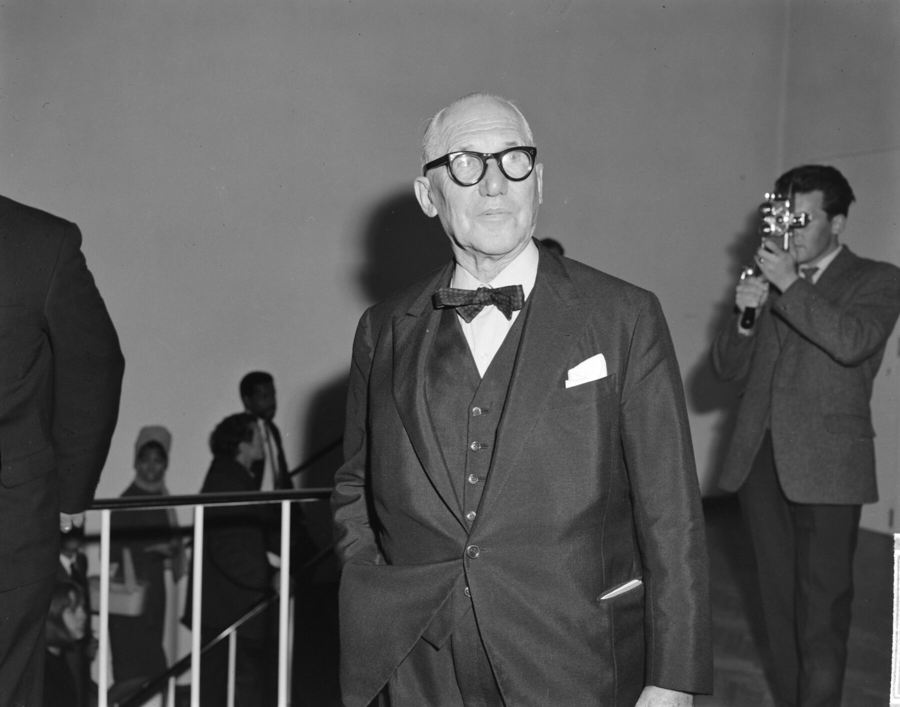 A man wearing black glasses and a dark suit looks at the camera. In the background, a man holds a video camera: the lens is directed towards the man wearing glasses.