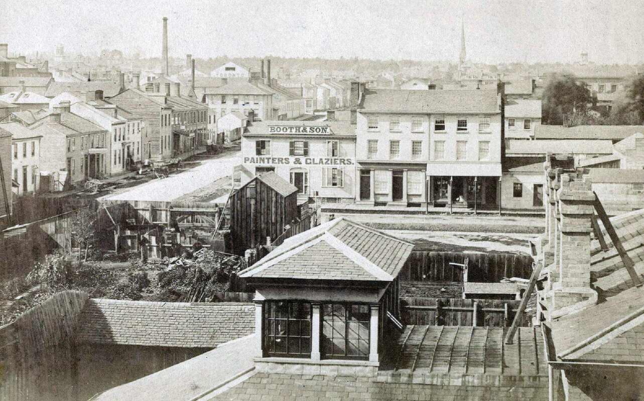 Historical photograph taken from a rooftop showing a collection of small buildings and houses, a portion of a street, and in the distance several church spires.
