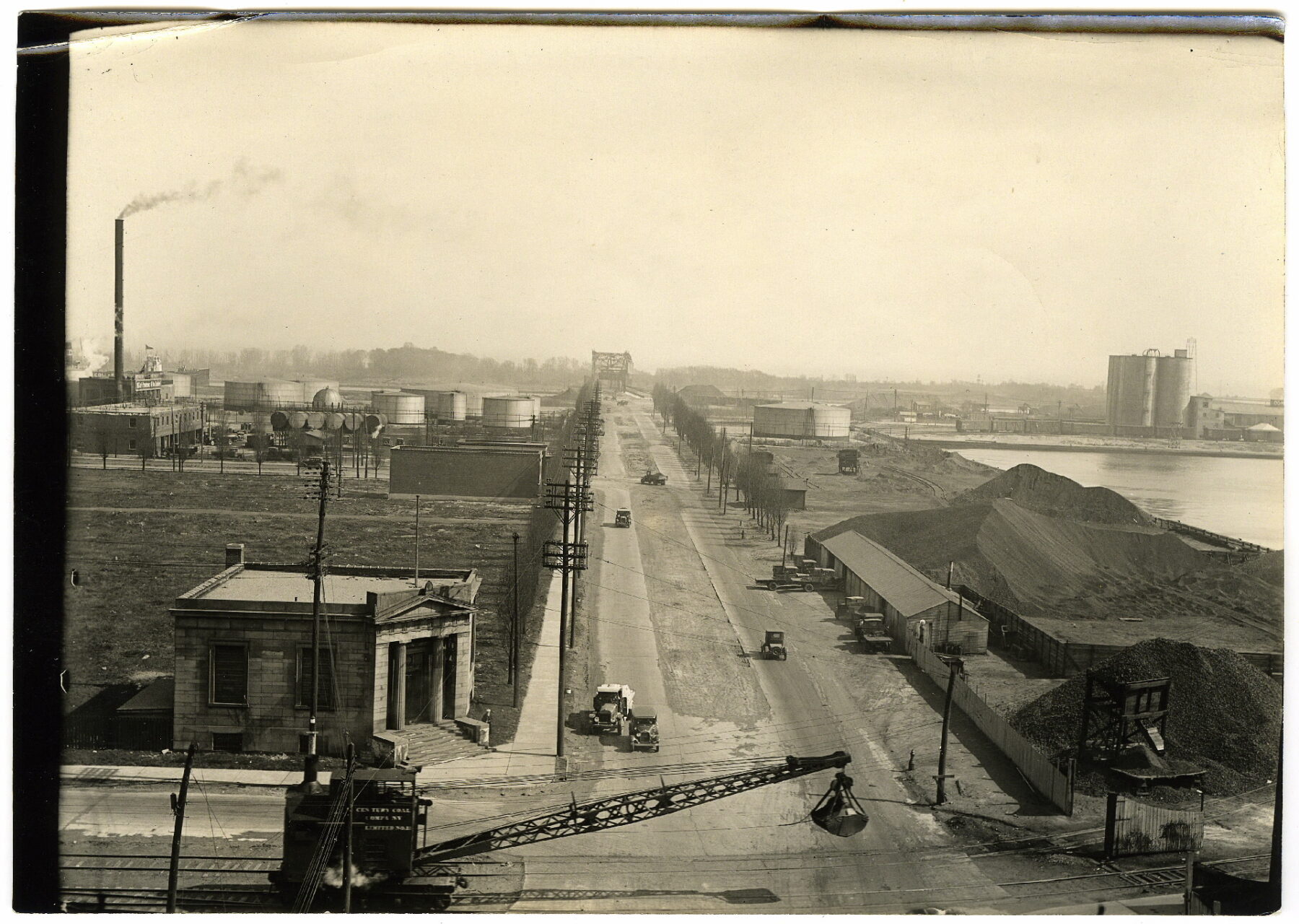 A black and white photograph that looks down on a road. The road is rough with a sizeable unpaved potion in the centre. Five cars are on the road, three going and two coming. They have a similar frame to a Ford Model T. A crane connected to a cart on rail tracks extends across the front of the image from left to right. There is a large building not far behind the crane. The entrance to the building has a classical-style portico with two large columns. This building is on the left side of the road. To the right are fenced-off yards containing mounds of dark material, likely coal. A few trees line the street to the right, while the electrical poles extend down its left side. A tall smokestack stands tall, closer to the top left of the picture. A small strand of smoke rises from it.