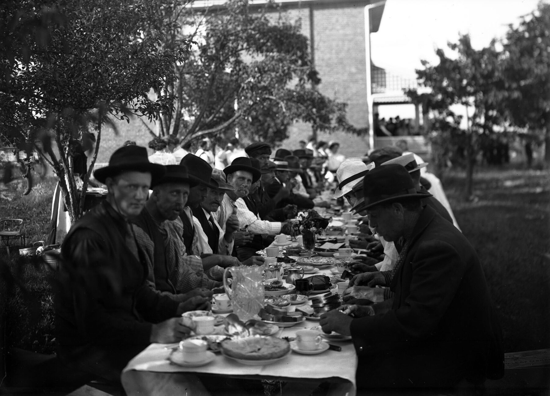 Black and white photo of a large group of men, all wearing hats, sitting at a long table with food, drinks, and various dishware on it. In the background is a large house, and off to the left of the photo is a tree. Behind the tree, a woman wearing a white apron is partly obscured from view. A few of the men look at the camera.