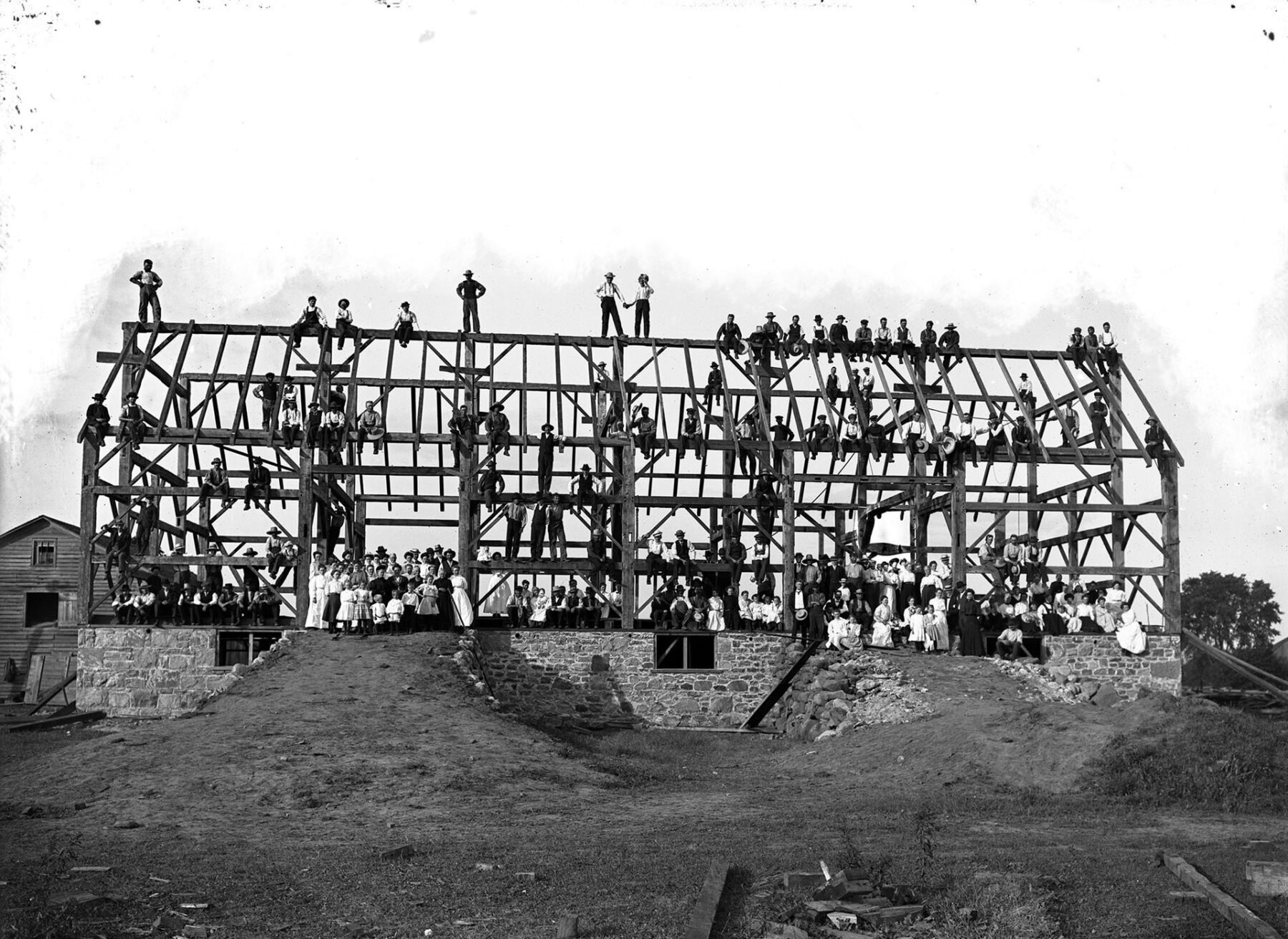 Black and white photo of a structure being built. Only the frame and stone foundation are completed. Many people pose for the photo, some of which are sitting or standing on the beams of the structure.