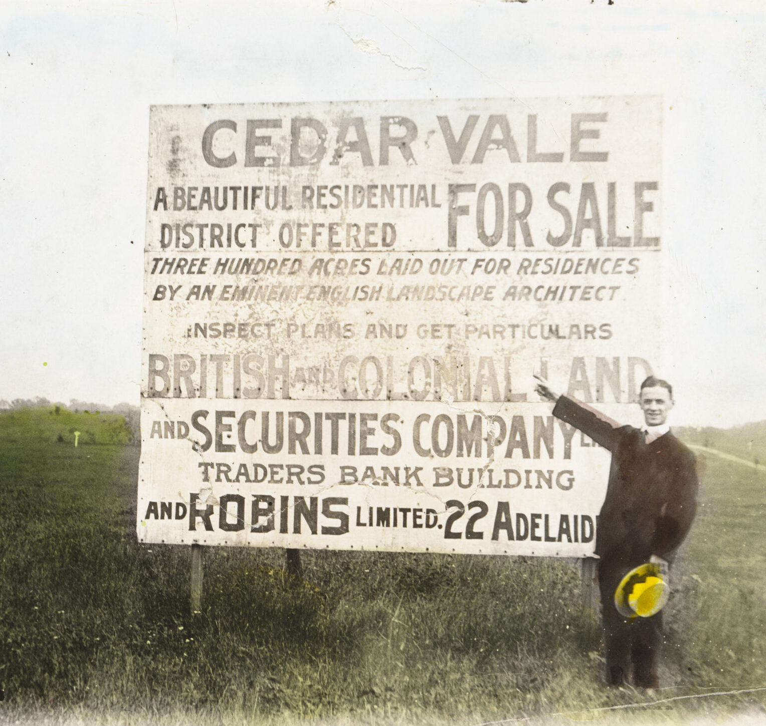 Image of Man standing in a cleared field and pointing at a large sign that reads CEDARVALE FOR SALE.