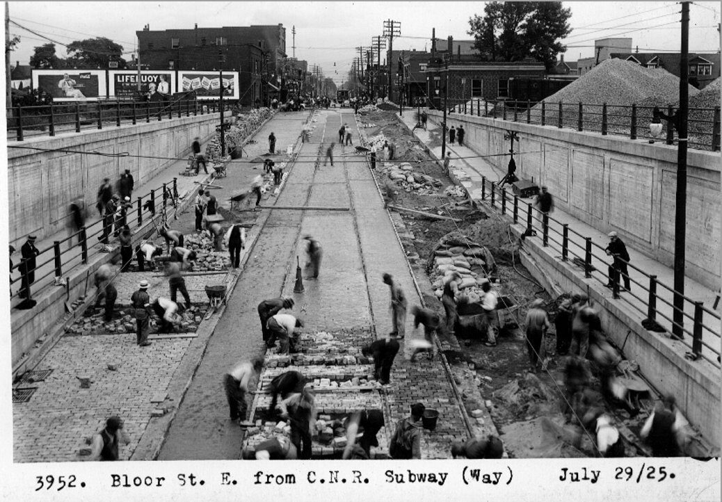Black and white photograph of many workers building an underpass on an existing city road. In the distance are trees and buildings.