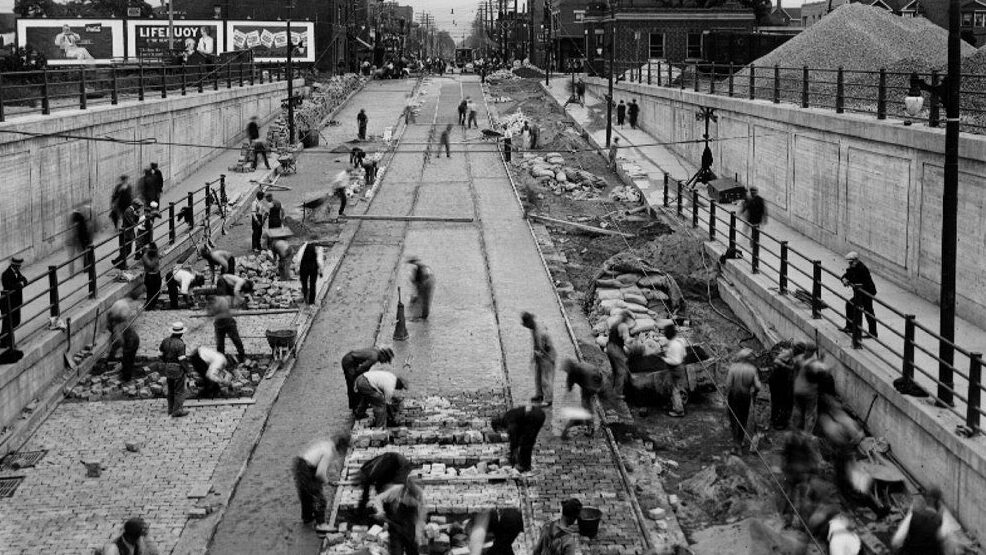 Black and white photograph of many workers building an underpass on an existing city road. In the distance are trees and buildings.