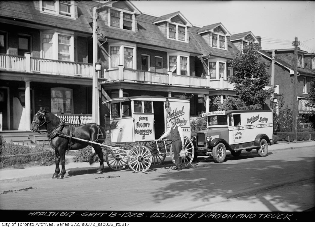 Black and white image of a milk delivery wagon and delivery van, both parked on a street. The milk man stands in front of the wagon and holds a milk crate with his left hand. A horse is harnessed to the wagon. Homes appear in the background with second-storey covered balconies.