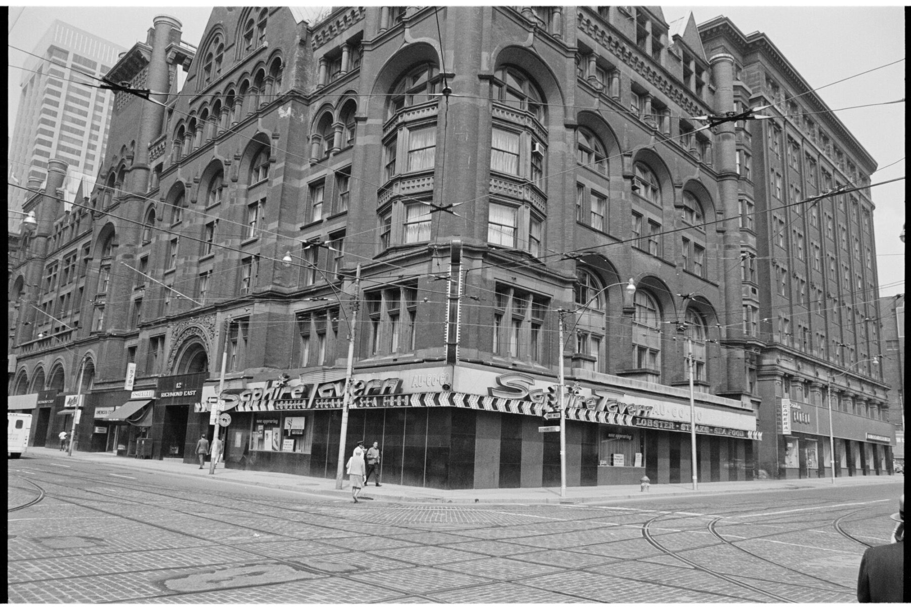 A black and white image depicts a brick building on a corner of the street, with a bulb light across the doorway that reads "The Saphire Tavern". The road is laid with streetcar tracks and wires criss-cross the air above.