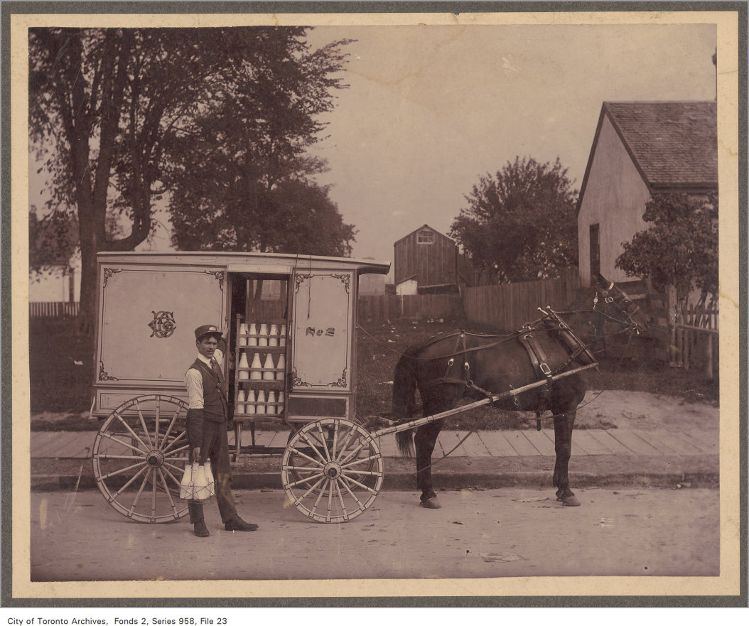 Sepia tone image of a horse-drawn milk cart. The milk man poses in front of the wagon, whose doors are open displaying the storage of milk bottles. He holds a crate of milk bottles with his right hand.